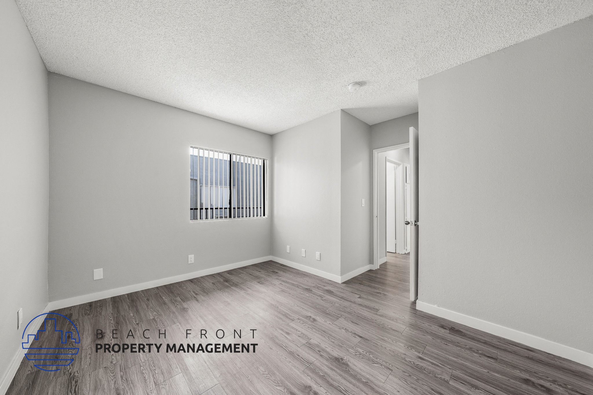 A spacious room with wooden flooring and a window with blinds.